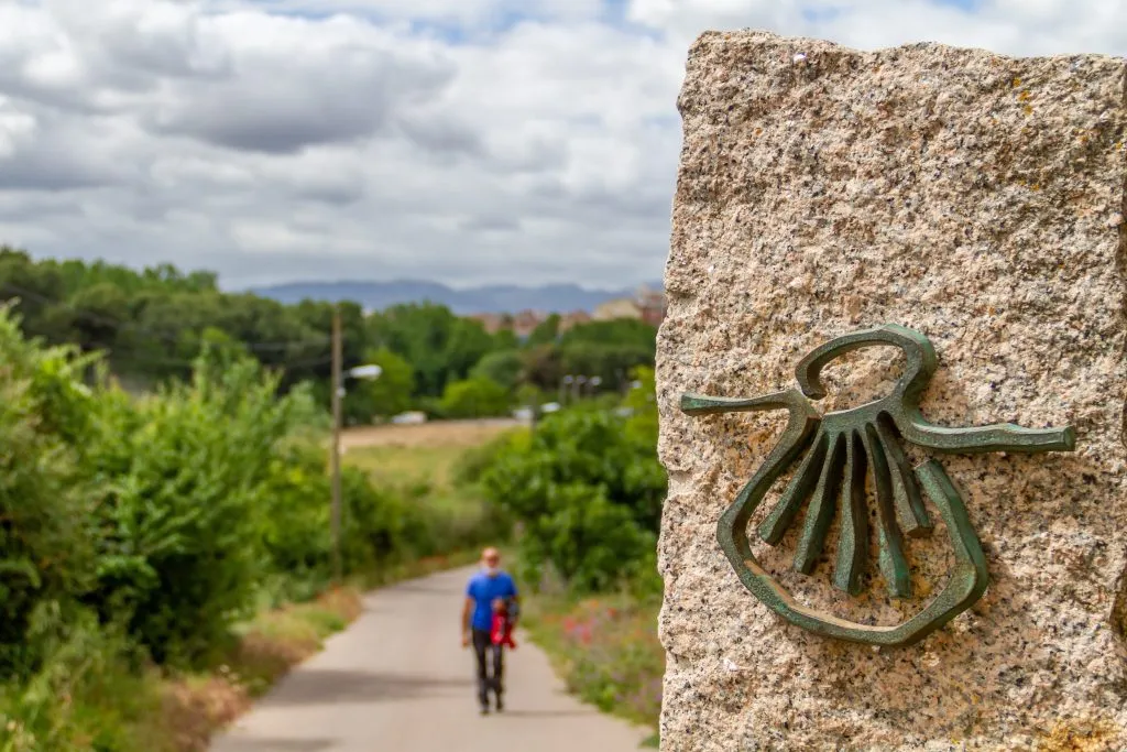 Pilgrim out of focus walks along the Camino de Santiago (Sant James Way) carrying a backpack next to a column on focus with the shell symbol of the Camino Santiago de Compostela