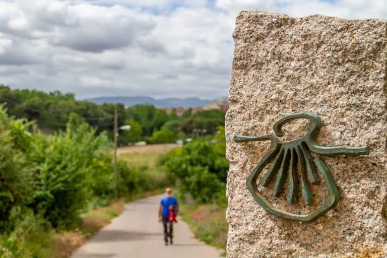 Pilgrim out of focus walks along the Camino de Santiago (Sant James Way) carrying a backpack next to a column on focus with the shell symbol of the Camino Santiago de Compostela